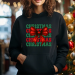 Black hoodie with green and red Christmas text and large red bow graphic, standing near decorated Christmas tree