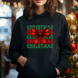 Black hoodie with green and red Christmas text and large red bow graphic, standing near decorated Christmas tree
