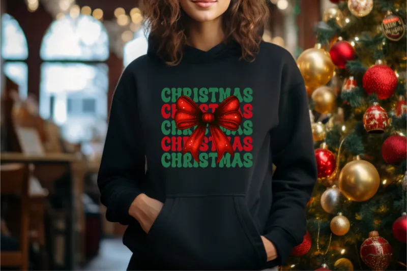 Black hoodie with green and red Christmas text and large red bow graphic, standing near decorated Christmas tree