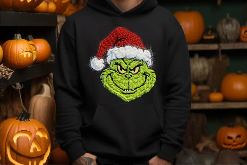 Green grinch character with santa hat printed on black hoodie surrounded by halloween pumpkins