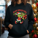 Black hoodie with In My Grinch Era Christmas design featuring cartoon Grinch in Santa outfit