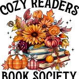The text reads "COZY READERS BOOK SOCIETY". The image depicts a stack of books surrounded by autumn-themed elements: sunflowers, pumpkins with bows, maple leaves, roses, and sparkling stars, creating a warm, inviting reading atmosphere.