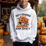 White hoodie with Cozy Readers Book Society design featuring autumn flowers, books, and pumpkins
