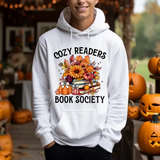 White hoodie with Cozy Readers Book Society design featuring autumn flowers, books, and pumpkins