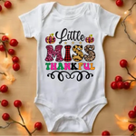 White baby onesie with Little Miss Thankful text in colorful leopard and plaid patterns, surrounded by red berries and fairy lights