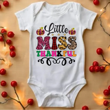 White baby onesie with Little Miss Thankful text in colorful leopard and plaid patterns, surrounded by red berries and fairy lights