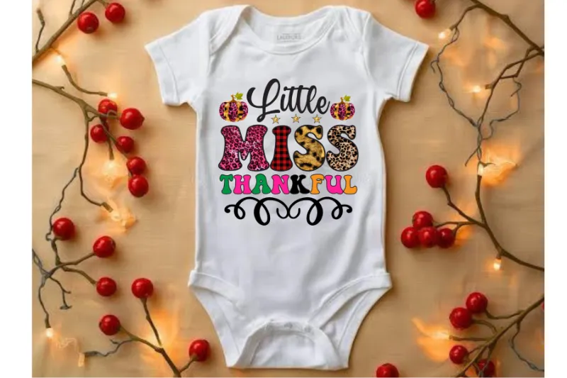 White baby onesie with Little Miss Thankful text in colorful leopard and plaid patterns, surrounded by red berries and fairy lights