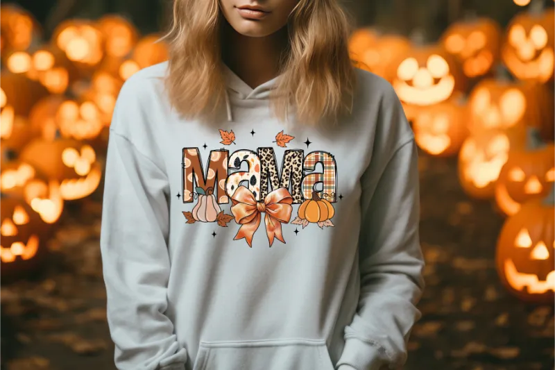 White hoodie with Mama text design featuring pumpkins, autumn leaves, and bow against jack-o-lantern background